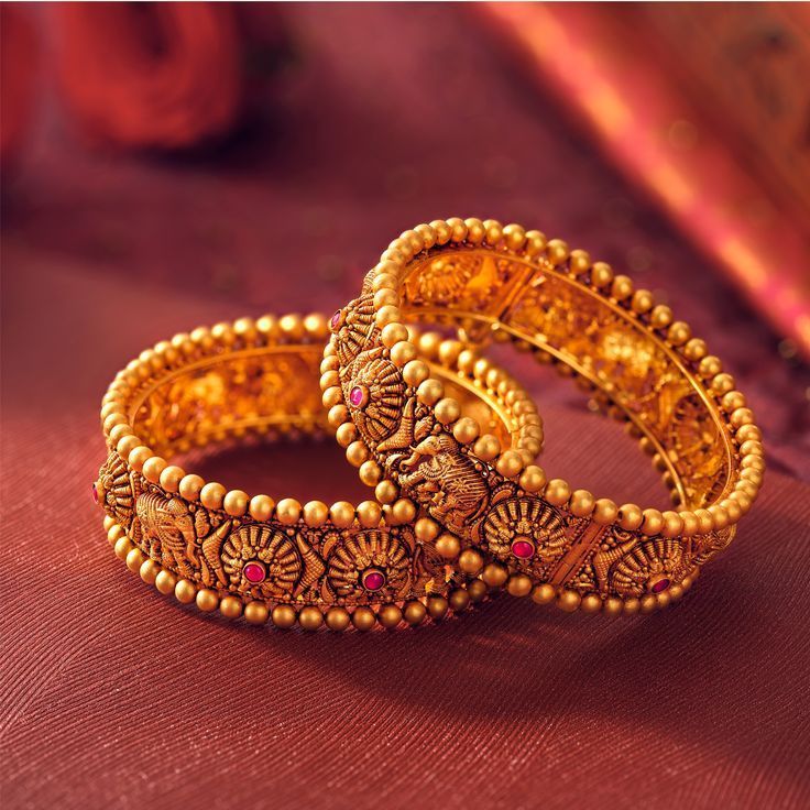 Two gold bangles with intricate designs on a red fabric background