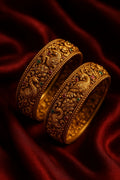 Two ornate gold bangles with intricate designs on a red fabric background