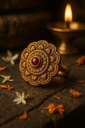 Decorative gold ring with a red gemstone on a dark surface with flowers and a lit candle in the background.
